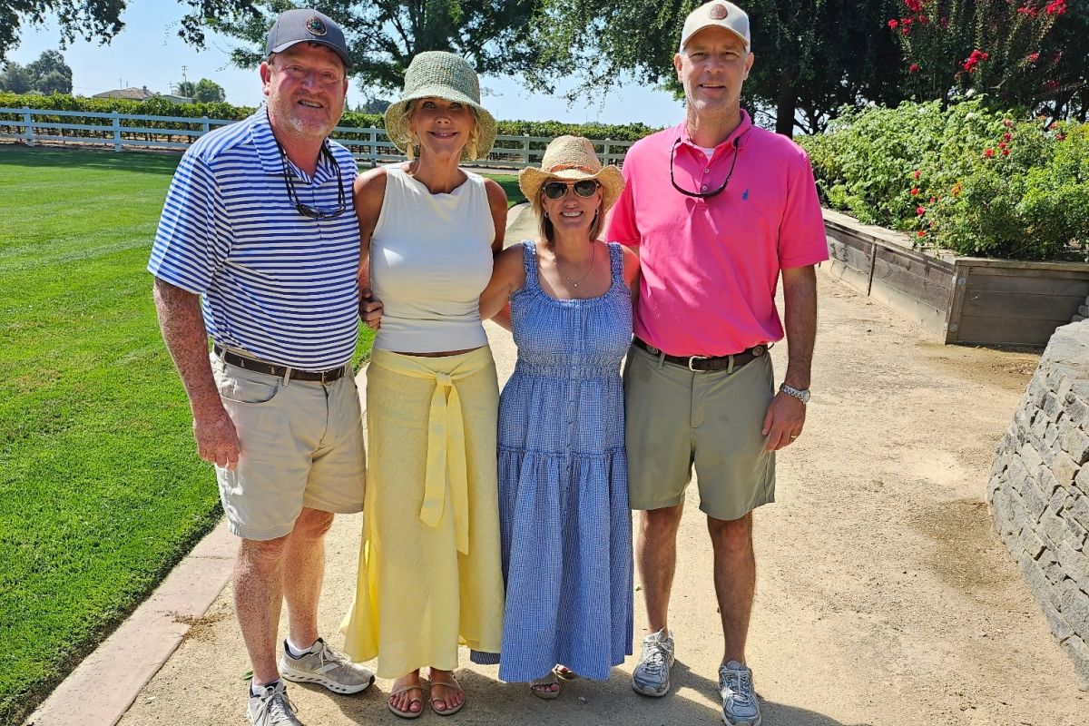 Four smiling wine tour guests standing together on a path beside a lawn and vineyard at an Edna Valley winery