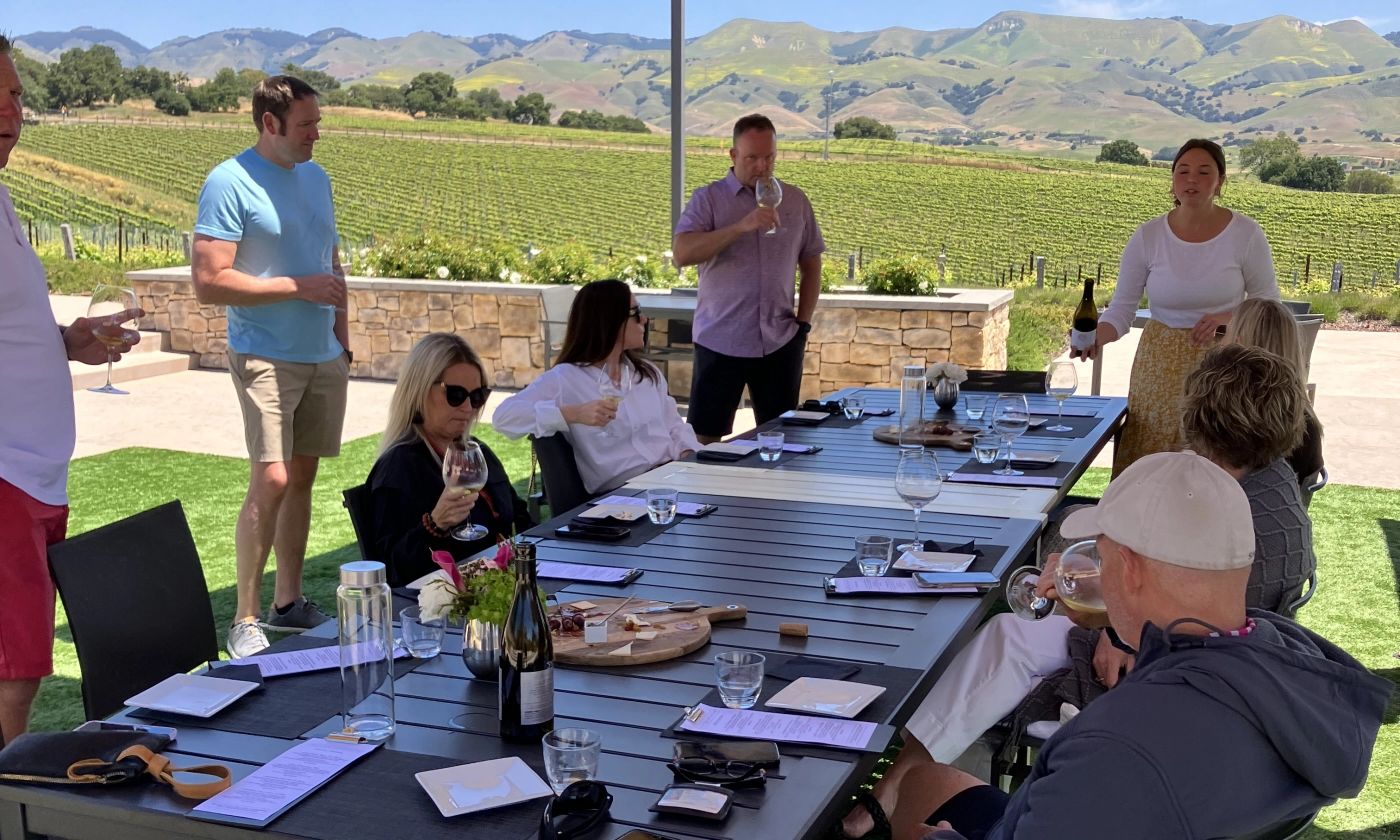 Wine tasting guests and a host pouring wine at a shaded table with views of vineyards and green hills in the SLO wine region