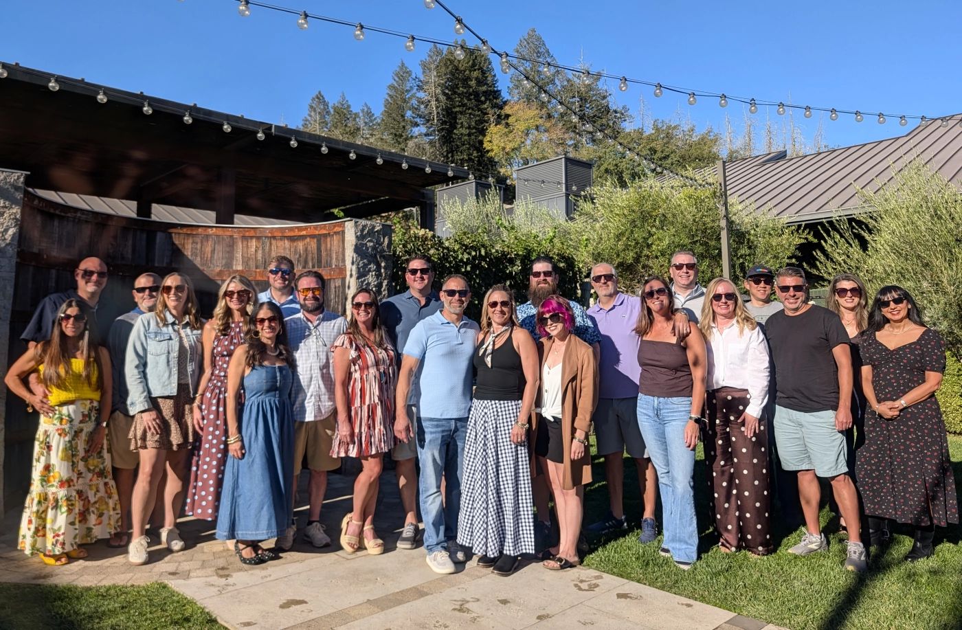Large group of wine tour guests in sunglasses smiling together under string lights at a winery patio